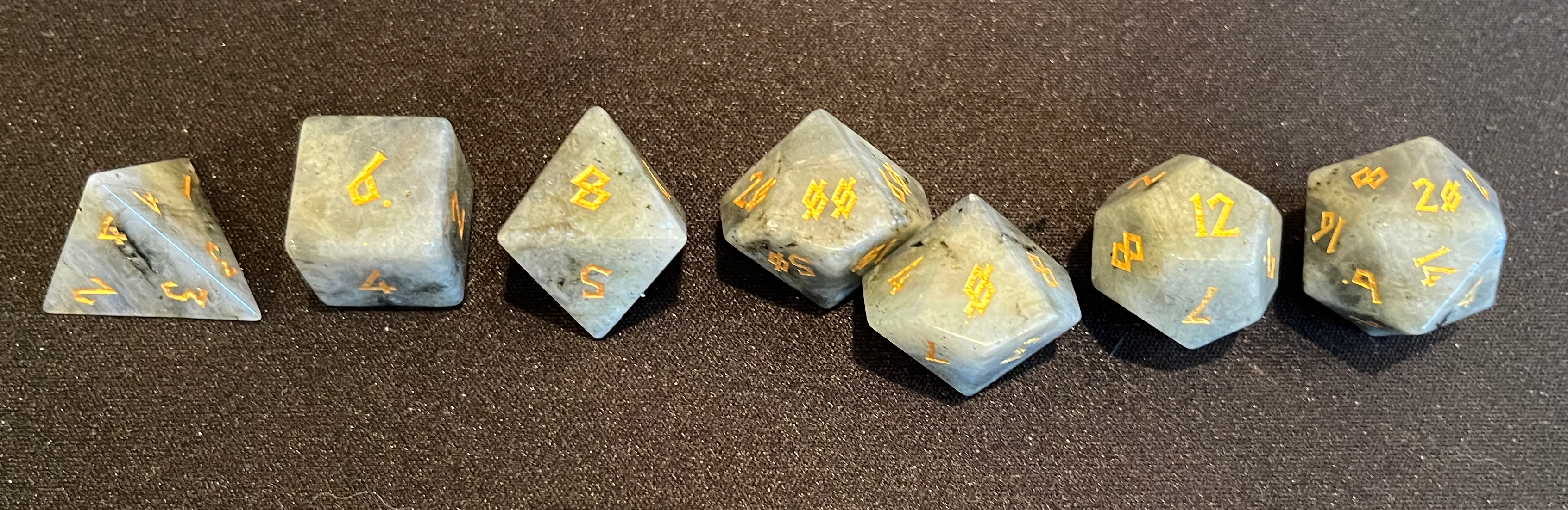 A set of 7 whitish-gray moonstone dice with gold-engraved Nordic-style font, from above.