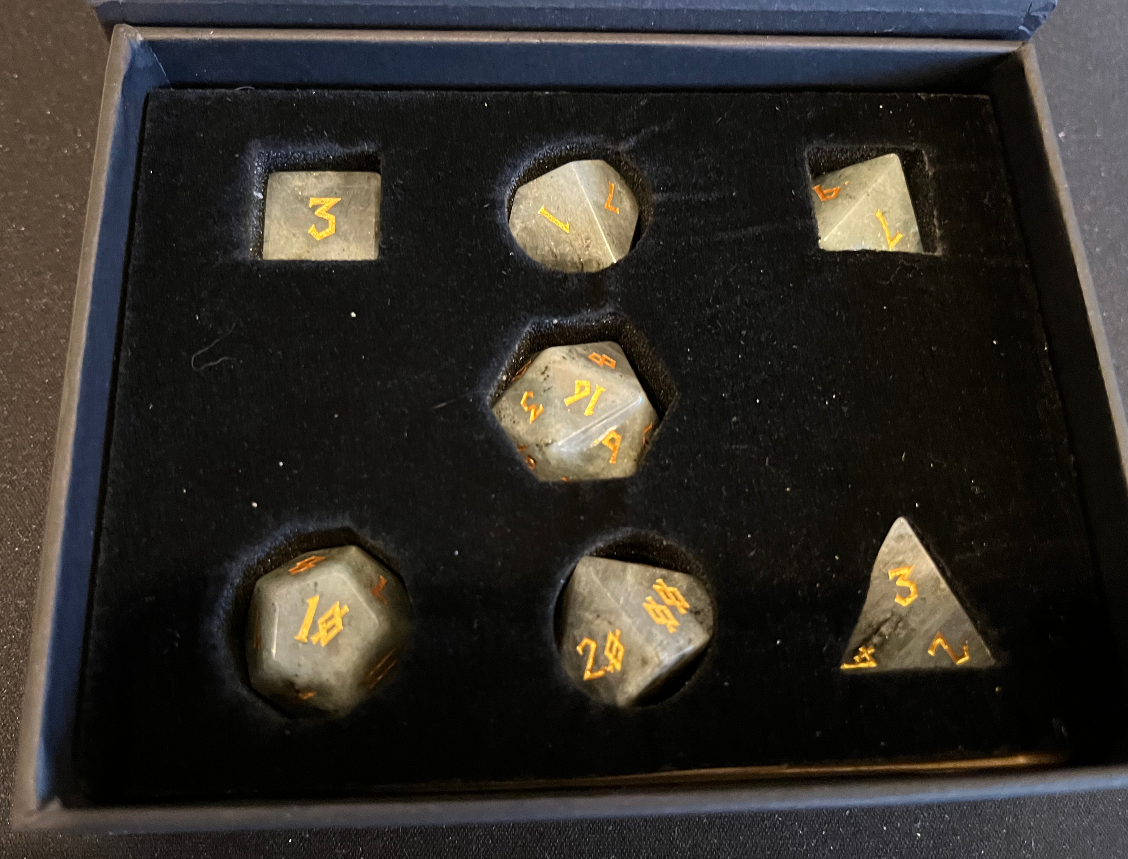 A set of 7 whitish-gray moonstone dice with gold-engraved Nordic-style font, from above.