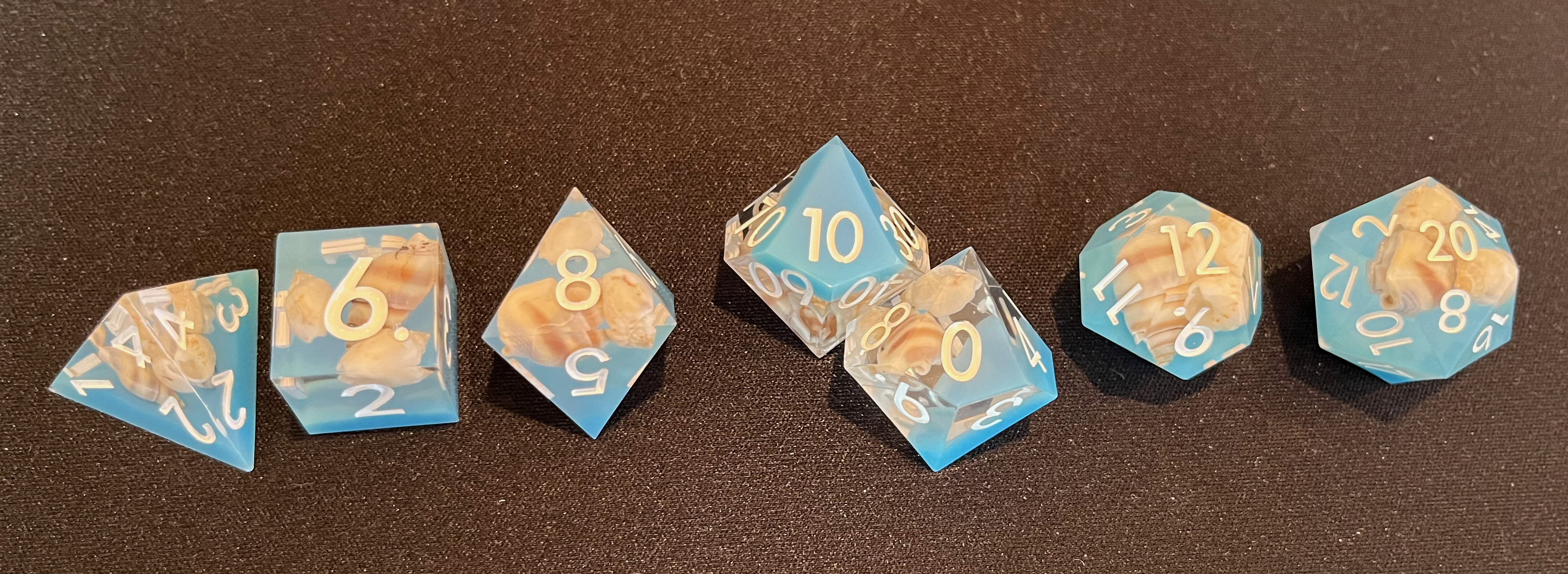 A set of 7 sharp-edged resin dice seen from above, partly transparent, partly translucent-to-opaque with an ocean blue color, and partly filled with tiny white seashells. They are inked with bright, easily readable white font.